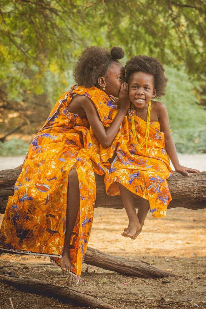 Two African children sitting on a log, sharing a secret in vibrant orange dresses outdoors.